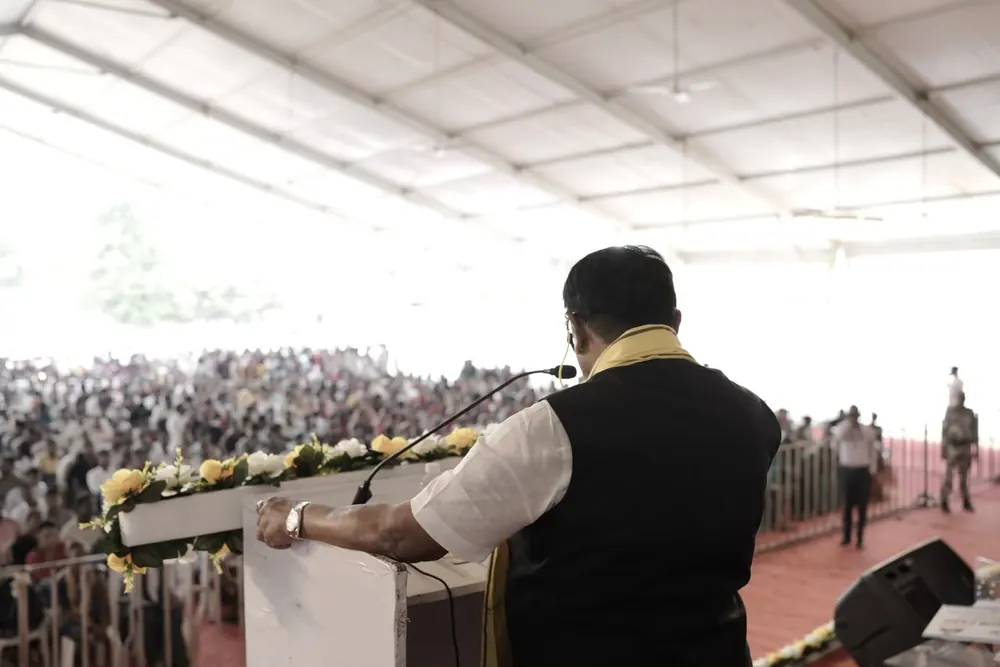 A speaker at a podium addressing a large crowd under a tent, seen from behind
