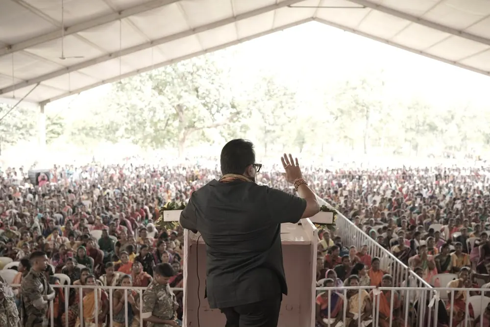 A speaker raising a hand to a vast crowd under a tent, seen from behind