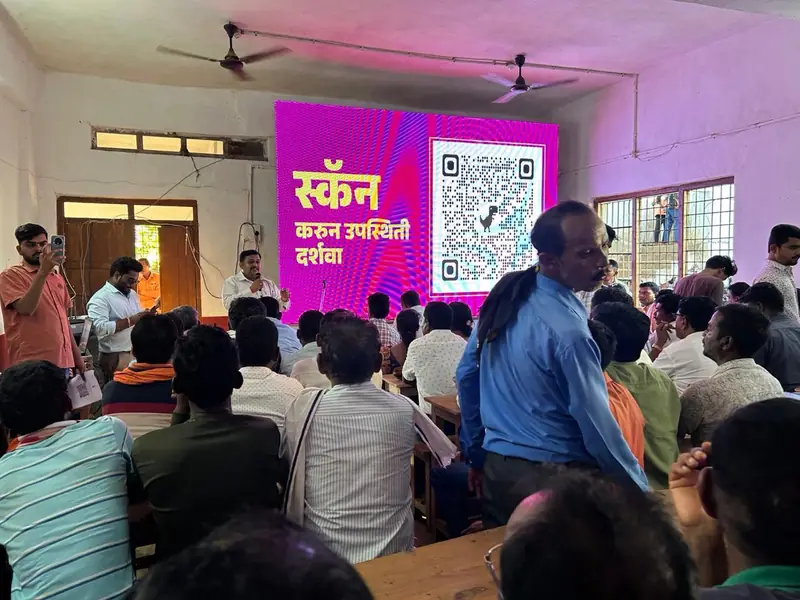 A speaker addressing a seated audience in a classroom beside a large screen with a QR code