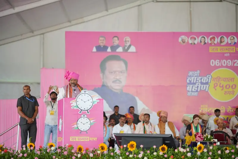 A speaker at a podium on a stage under a tent, with leaders seated below a large banner